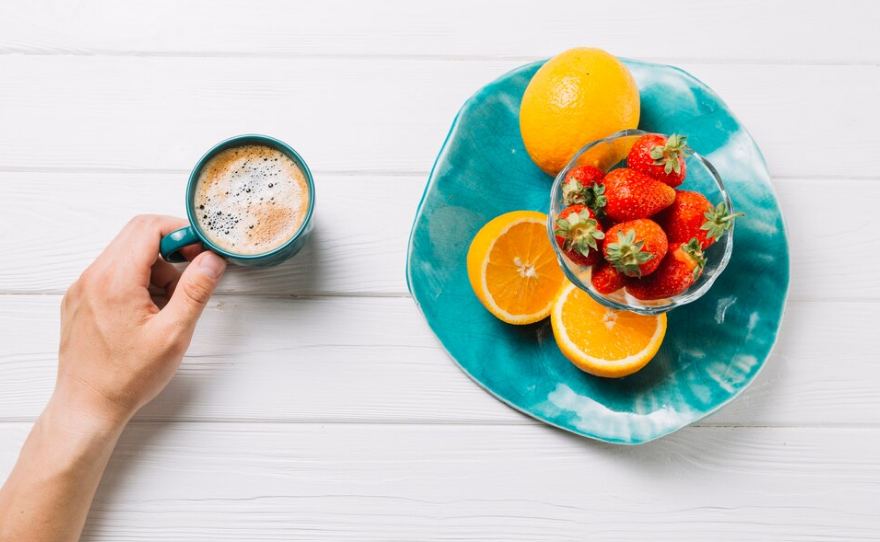 Mano sosteniendo una taza de café junto a un plato con fresas y naranjas sobre una mesa