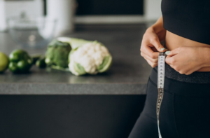 Mujer tomando sus medidas de cintura en la cocina con vegetales en el fondo.