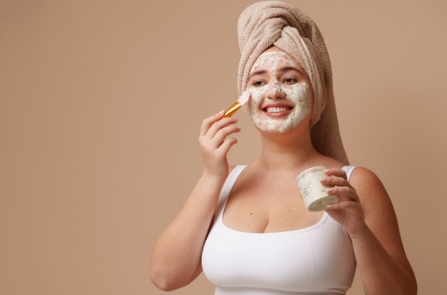 [Una mujer joven con una toalla en la cabeza aplicando una mascarilla de arcilla blanca en su rostro]