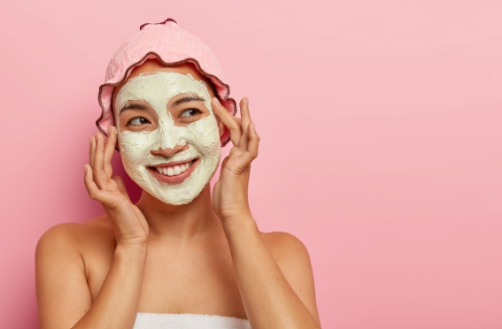 [Mujer sonriente con una mascarilla de arcilla blanca y un gorro de ducha rosa]