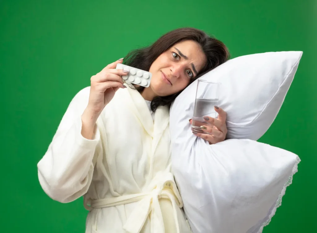 Mujer con bata sosteniendo una almohada, un vaso de agua y un blíster de pastillas mientras muestra cansancio por dificultad para dormir